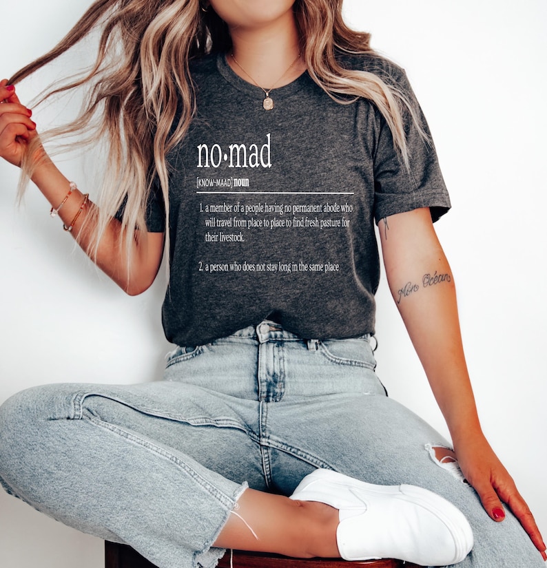 May include: A dark gray t-shirt with a white graphic design that reads "no-mad" and defines the word as a person who does not stay long in the same place.