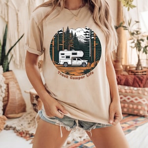 May include: Beige t-shirt featuring a circular graphic of a white truck camper in a forest scene with mountains and the text "Truck Camper Life" in brown. The shirt is a neutral color.