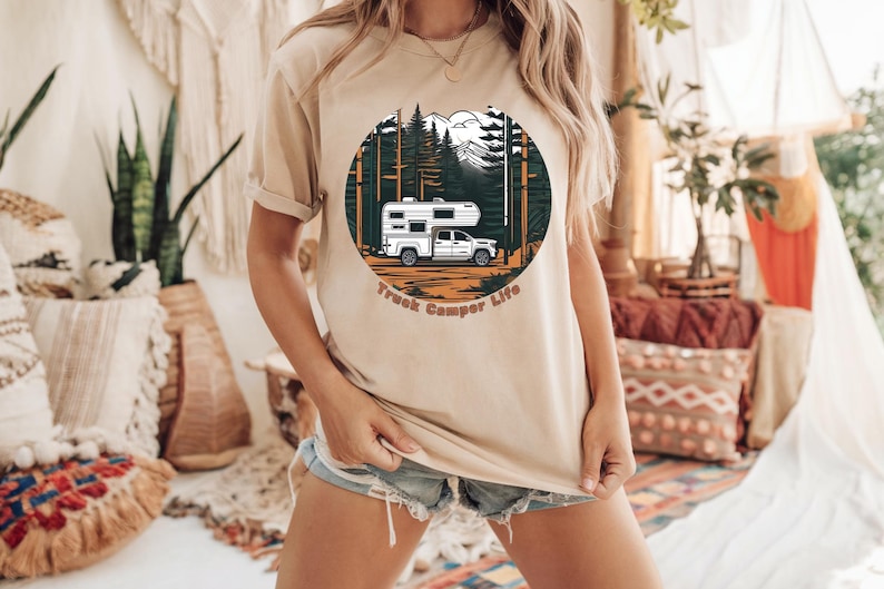 May include: Beige t-shirt featuring a circular graphic of a white truck camper in a forest scene with mountains and the text "Truck Camper Life" in brown. The shirt is a neutral color.