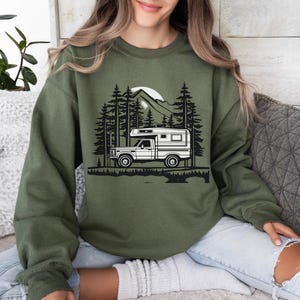 May include: Olive green sweatshirt featuring a black and white graphic of a camper truck in a forest scene with a mountain and sun. The design includes tall pine trees and a detailed landscape.
