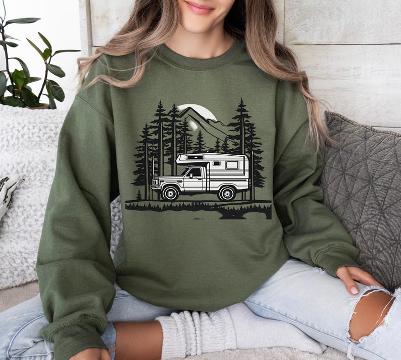 May include: Olive green sweatshirt featuring a black and white graphic of a camper truck in a forest scene with a mountain and sun. The design includes tall pine trees and a detailed landscape.