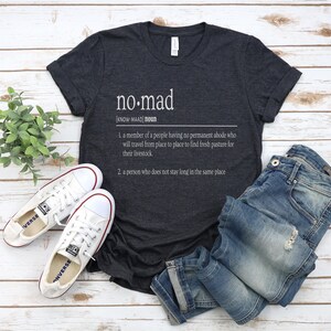 May include: A dark gray t-shirt with the word "no-mad" defined as a person who does not stay long in the same place. The shirt is laid out on a white wooden surface with a pair of white canvas shoes and blue jeans.