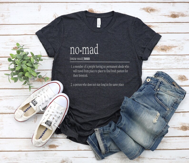 May include: A dark gray t-shirt with the word "no-mad" defined as a person who does not stay long in the same place. The shirt is laid out on a white wooden surface with a pair of white canvas shoes and blue jeans.