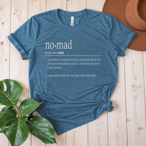 May include: A teal blue t-shirt with a white graphic design that defines the word "no-mad" as a person who does not stay long in the same place.