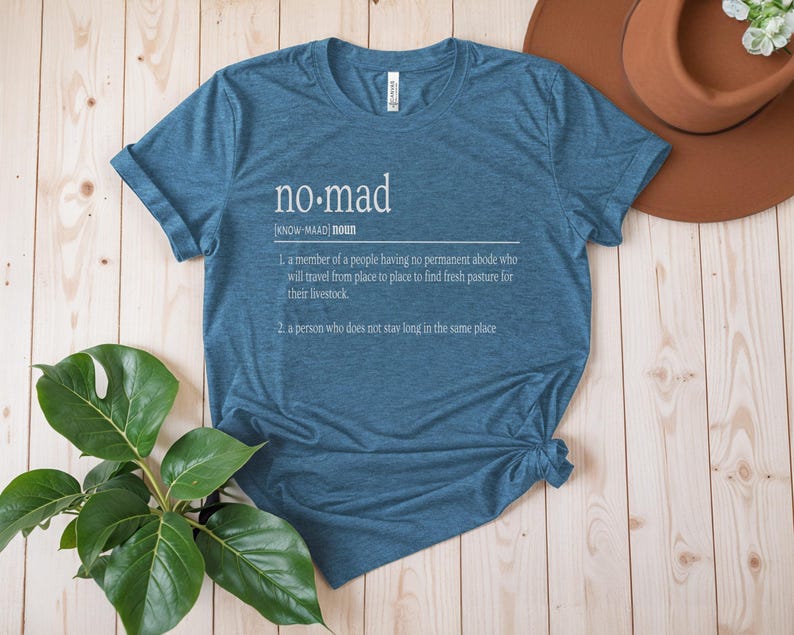 May include: A teal blue t-shirt with a white graphic design that defines the word "no-mad" as a person who does not stay long in the same place.