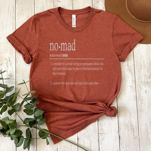 May include: A rust-colored t-shirt with the word "no-mad" defined as a person who does not stay long in the same place.