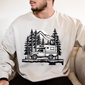 May include: Cream-colored sweatshirt featuring a black and white graphic of a camper truck in a forest setting with mountains and trees. The design evokes a sense of adventure and outdoor travel.