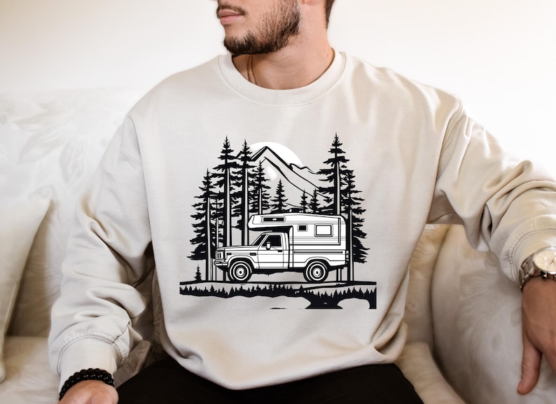 May include: Cream-colored sweatshirt featuring a black and white graphic of a camper truck in a forest setting with mountains and trees. The design evokes a sense of adventure and outdoor travel.