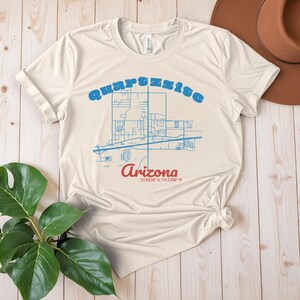 May include: A beige t-shirt featuring a blue outline of a map of Quartzsite, Arizona. The text "Quartzsite, Arizona 33.8339&deg; N 114.2289&deg; W" is printed on the shirt.