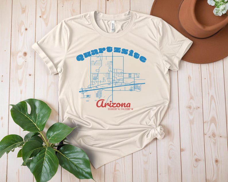 May include: A beige t-shirt featuring a blue outline of a map of Quartzsite, Arizona. The text "Quartzsite, Arizona 33.8339&deg; N 114.2289&deg; W" is printed on the shirt.