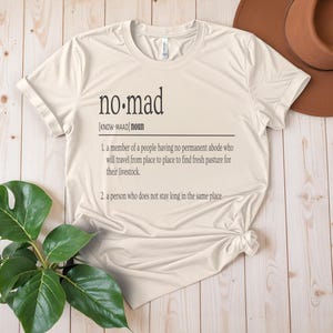 May include: A beige t-shirt with the word "no-mad" defined as a person who does not stay long in the same place. The definition is written in black text on the shirt.
