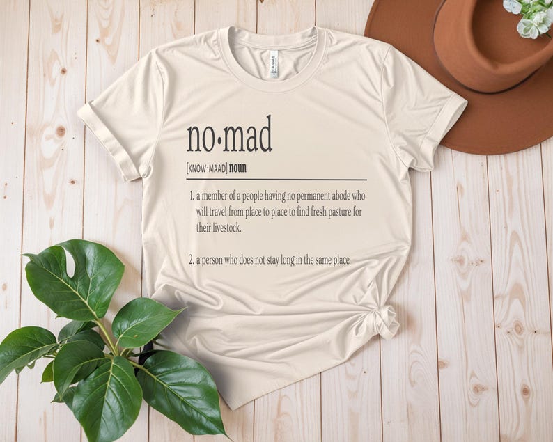 May include: A beige t-shirt with the word "no-mad" defined as a person who does not stay long in the same place. The definition is written in black text on the shirt.