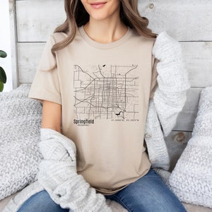 Pode incluir: Uma camiseta bege com um contorno preto e branco de um mapa da cidade. O mapa é rotulado como "Springfield - Missouri" e inclui as coordenadas "37.2090° N, 93.2923° W".