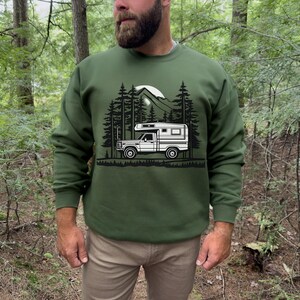 May include: Green sweatshirt featuring a black and white graphic of a camper van in a forest setting. The design includes trees, mountains, and a full moon. The sweatshirt is a solid green color.