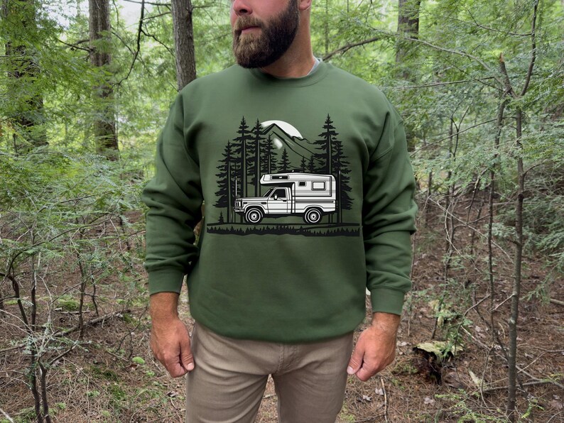 May include: Green sweatshirt featuring a black and white graphic of a camper van in a forest setting. The design includes trees, mountains, and a full moon. The sweatshirt is a solid green color.