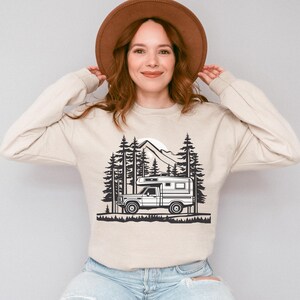 May include: Beige long-sleeve sweatshirt featuring a black and white graphic of a camper van in a forest scene with mountains and trees. The sweatshirt is made of a soft material and has a crew neck.