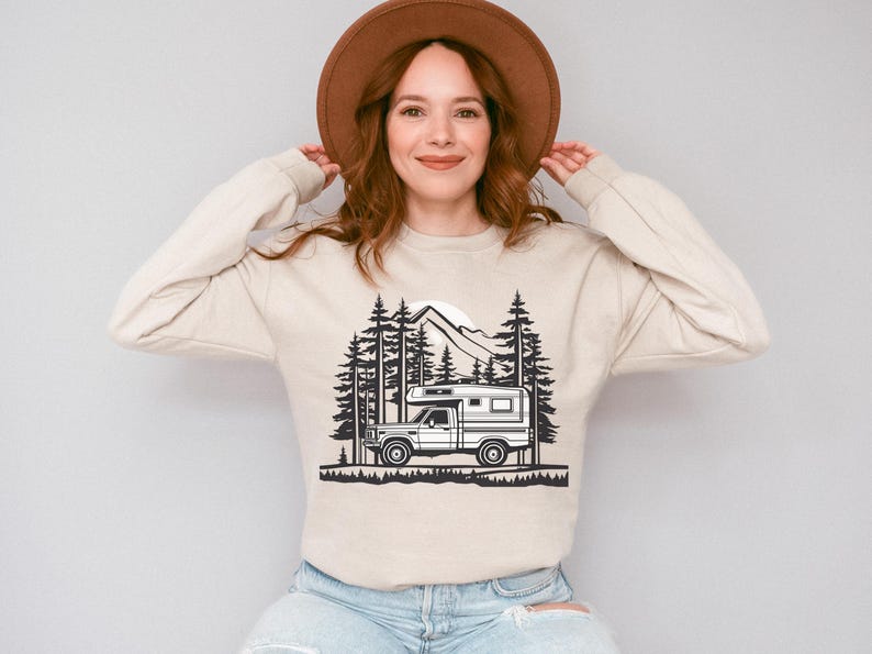 May include: Beige long-sleeve sweatshirt featuring a black and white graphic of a camper van in a forest scene with mountains and trees. The sweatshirt is made of a soft material and has a crew neck.