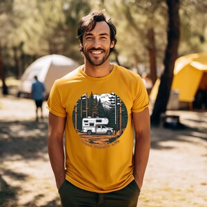 May include: Mustard yellow t-shirt featuring a circular graphic of a white camper truck in a forest scene with mountains. The text "Truck Camper Life" is below the graphic. The person is wearing dark green pants.