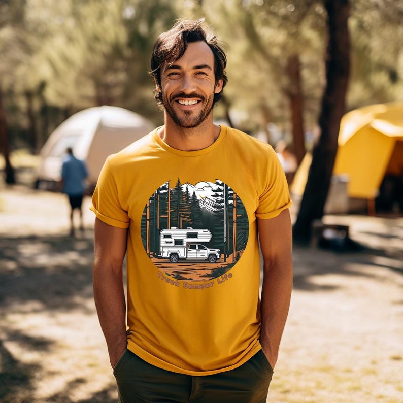May include: Mustard yellow t-shirt featuring a circular graphic of a white camper truck in a forest scene with mountains. The text "Truck Camper Life" is below the graphic. The person is wearing dark green pants.