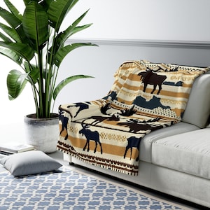 Puede incluir: Una manta de color crema con un patrón de alces marrones, negros y azules. La manta está doblada y colocada sobre un sofá blanco.