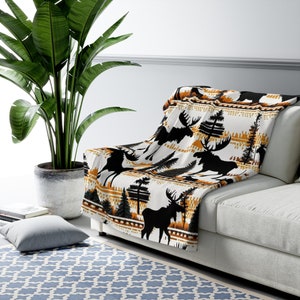 Puede incluir: Una manta de tiro blanca, negra y naranja con un patrón repetitivo de alces y pinos. La manta está drapeada sobre un sofá blanco.