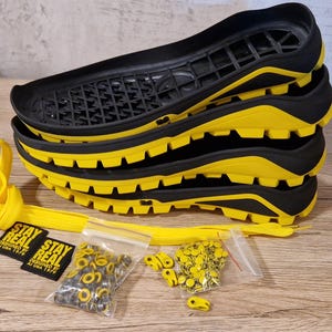 Könnte beinhalten: Ein Stapel schwarzer und gelber Schuhsohlen mit passenden gelben Schnürsenkeln, Ösen und Etiketten mit der Aufschrift "STAY REAL". Die Sohlen haben ein robustes Profildesign. Zusätzliche Hardware ist enthalten.