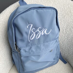 May include: A pale blue backpack featuring the name "Issa" embroidered in white cursive. The bag has a front pocket, a top handle, and adjustable shoulder straps. A silver zip and a small silver metal detail are visible.
