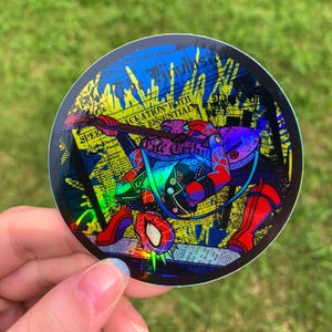 May include: A circular holographic sticker featuring a stylized character in a red and blue suit playing a guitar. The background consists of yellow and blue newspaper clippings with visible text including The Hindu and Evacuation Both Essential.