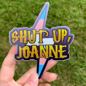 May include: A die-cut sticker featuring a lightning bolt shape with a light blue, pink, and white striped background. The text SHUT UP, JOANNE is printed in a bold, glittery gold font with a dark purple outline, creating a high-contrast design.