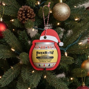 May include: A Christmas tree decorated with ornaments and lights. A red Santa hat-shaped ornament frames a BuzzBall Cocktails can, labeled "Holiday Nog." Other ornaments include red and gold baubles and a pinecone.