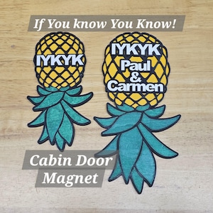 May include: Two yellow pineapple-shaped cabin door magnets with the text "IYKYK" on one and "IYKYK Paul & Carmen" on the other. The pineapples have green leaves and are on a wooden surface.