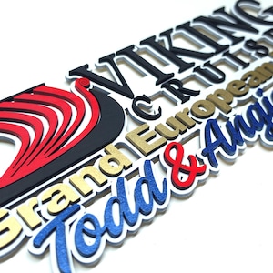 May include: A multi-layered, three-dimensional sign featuring the text VIKING CRUISES, Grand European Tour, and Todd & Angie 2024. The design includes a black and red stylized ship logo, with lettering in black, gold, red, and blue on a white base.