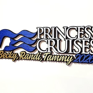 Op de afbeelding: Een houten bord met de woorden "PRINCESS CRUISES" in witte en zwarte letters. Het Princess Cruises-logo is blauw. Daaronder staan de namen "Becky, Randi, Tammy" in goud en het jaar "2026" in blauw.
