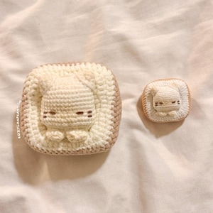 May include: Two handmade crochet cat-shaped items. The larger one is a square with a beige cat in the center, surrounded by a white border and a brown edge. The smaller one is a miniature version of the same design. The text "COZYLOAFCAT" is visible on the larger item.