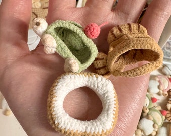 May include: A hand holding three crocheted toys: a green turtle with a pink flower, a brown and tan hat, and a white and tan ring.