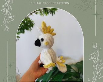 May include: A crocheted cockatoo amigurumi toy with a white body, yellow crest, and black eyes. The toy is being held in a hand against a green background.
