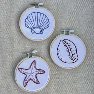 May include: Three embroidered hoops featuring sea life designs. One hoop has a blue seashell, another has a brown cowrie shell, and the third has a red starfish. The designs are on light blue fabric, set in wooden hoops.