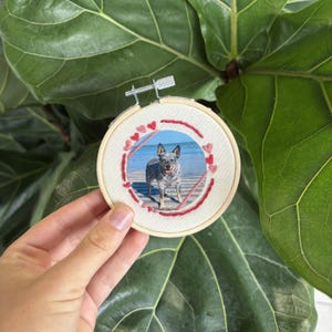 Könnte beinhalten: Ein runder Stickrahmen mit einem Foto eines Hundes am Strand, umgeben von roten Herzstickereien. Der Stickrahmen wird vor einem großen grünen Blatt gehalten.
