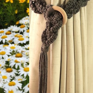 May include: A gray macrame curtain tieback with a wooden ring. The tieback is wrapped around a beige curtain. The macrame has a tassel at the end. Daisies and yellow flowers are in the background.