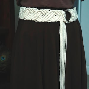 May include: Cream-colored macrame belt with a geometric design and a metal ring closure. The belt features a long fringe detail hanging down. The belt is worn over a dark brown skirt and a brown top.