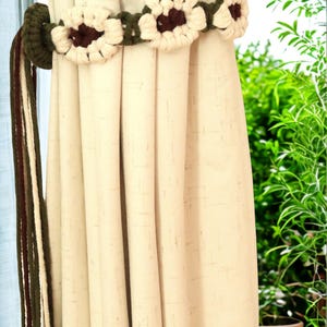 May include: Cream-colored curtains are held back by a crocheted tieback. The tieback features a series of white and brown flower-shaped accents on a green band. A matching green and brown rope hangs to the left of the curtains. The background shows green plants.