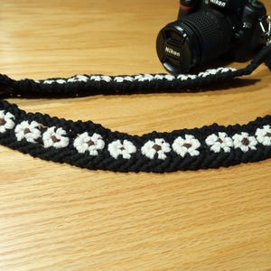 May include: Black and white daisy-patterned camera strap. The strap is made of a woven material with white daisy-shaped flowers and brown centers. A black Nikon camera is attached to the strap.