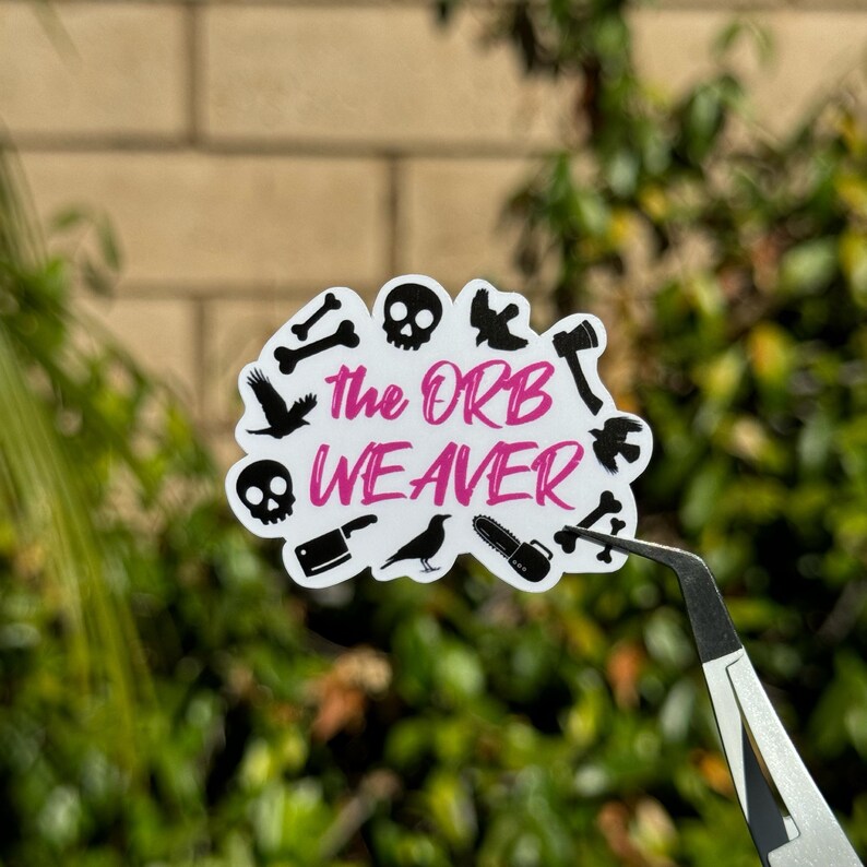 May include: A white sticker with black and pink text that reads "The Orb Weaver". The sticker is surrounded by black silhouette images of bones, crows, a skull, a chainsaw, an axe, and a knife.