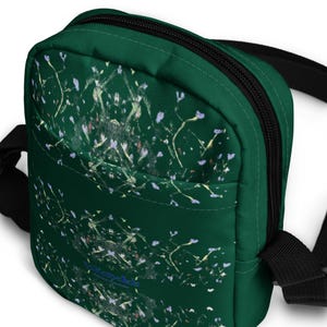 May include: A green canvas crossbody bag with a black strap and a white and blue abstract pattern. The bag has a zipper closure and a small pocket on the front.