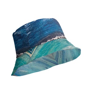 May include: Bucket hat with an abstract ocean-themed design. The hat features a dark blue sky, a white shoreline, and turquoise water. The brim and crown are painted with visible brushstrokes, creating a textured appearance. The hat is a stylish accessory.