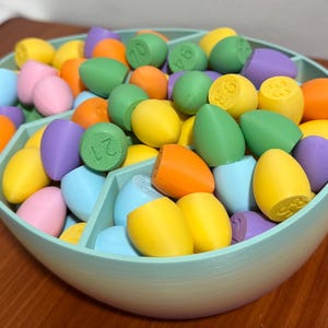 May include: A pastel-colored bowl filled with colorful plastic eggs, each with a number printed on it. The bowl is divided into sections and has a rainbow gradient.