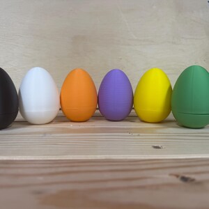 May include: Six brightly coloured, egg-shaped containers. The colours are black, white, orange, purple, yellow, and green. The containers are arranged on a wooden surface.