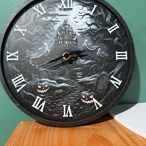 May include: Black round wall clock with a Halloween theme. The clock face features a spooky haunted house, ghosts, pumpkins, and tombstones. White Roman numerals mark the hours. The clock hands are black with a gold center.