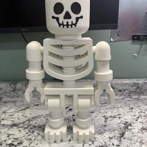 May include: A white, 3D-printed skeleton figure, resembling a large toy block. The head features a black skull design. The figure has articulated arms and legs, and a rib cage design. The figure is standing on a speckled surface.