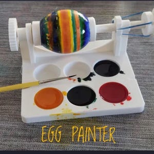 May include: A white plastic egg painting station with a colorful painted egg secured in the center. A paintbrush rests on a white palette with various paint colors. The text "EGG PAINTER" is written in yellow.